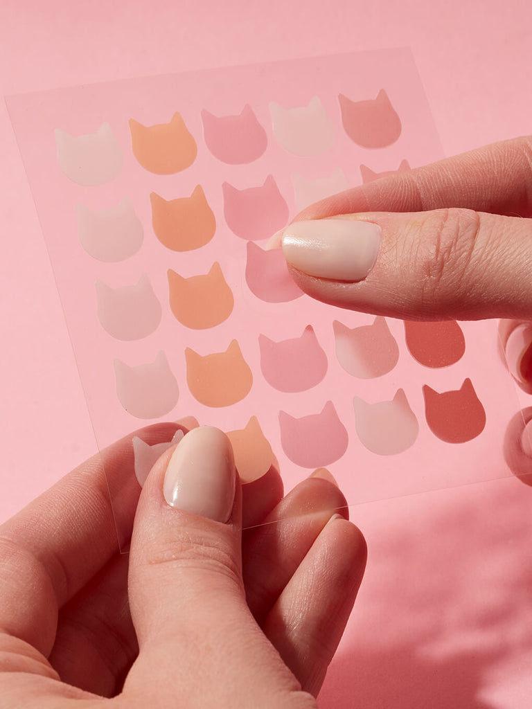 hand holding sheet of cat shaped pimple patches on pink background