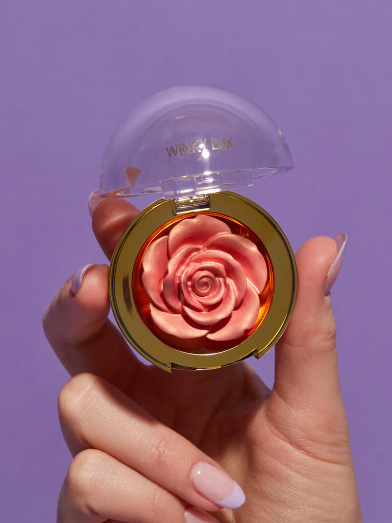 Rosé -- hand holding up cheeky rose cream highlighter on purple background
