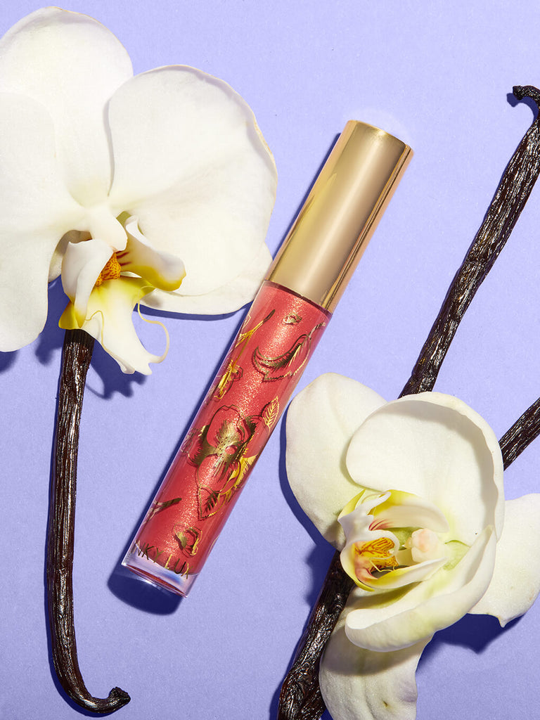 Meow -- glossy boss lip gloss flat lay on purple background surrounded by orchids